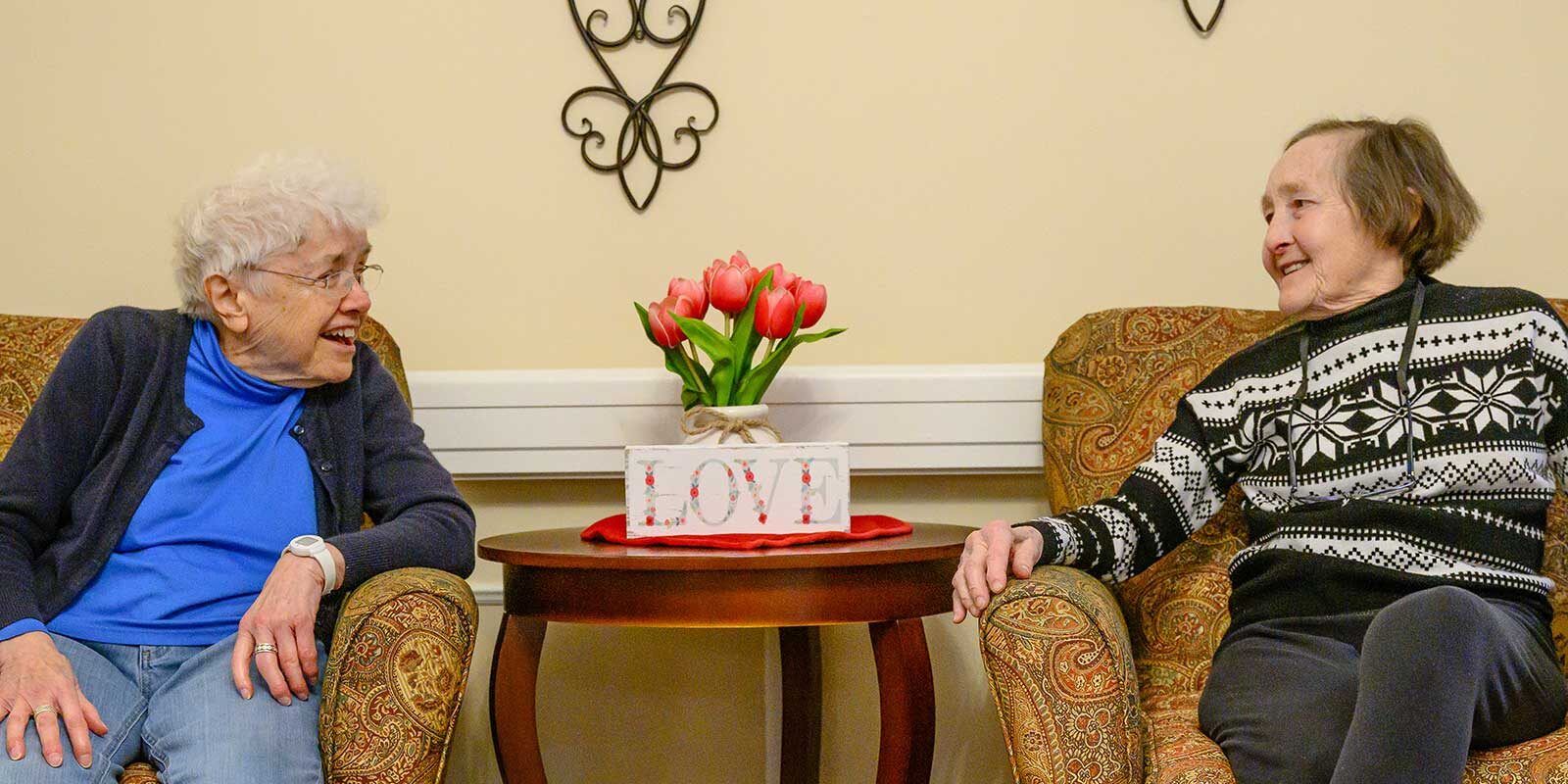 Two elderly women smiling and chatting in cozy chairs.